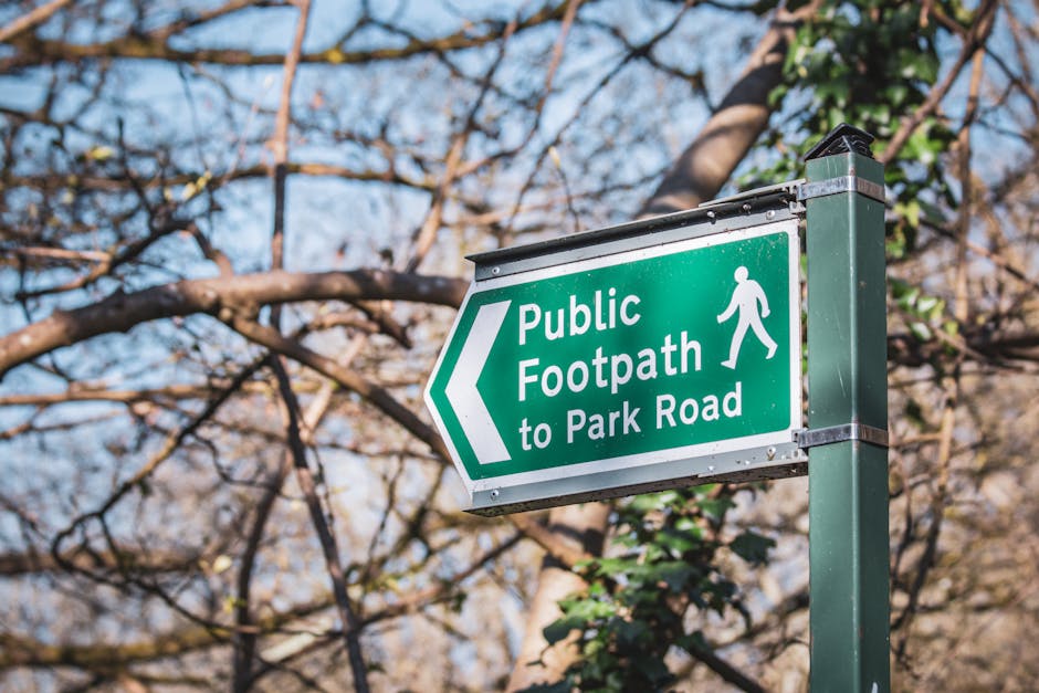 A green rectangular directional sign mounted on a dark green metal pole, indicating a pedestrian footpath leading to Park Road. The sign features white text reading 'Public Footpath to Park Road' with